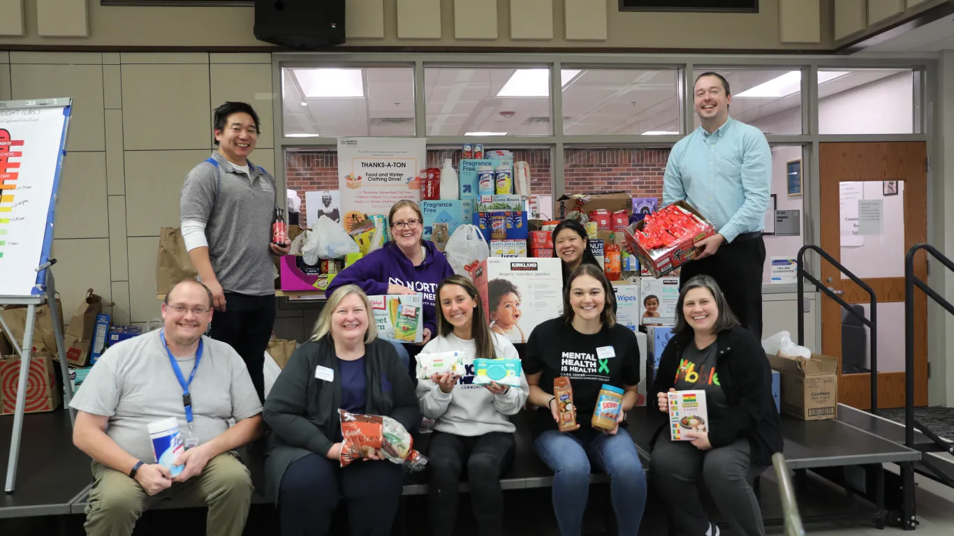 a group photo of NHCC staff smiling with food donations in the Campus Center during their Food Drive event. 