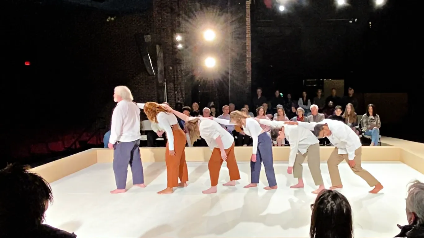 The chorus members of Antigone standing one behind the other with their arms stretched out. All on stage.