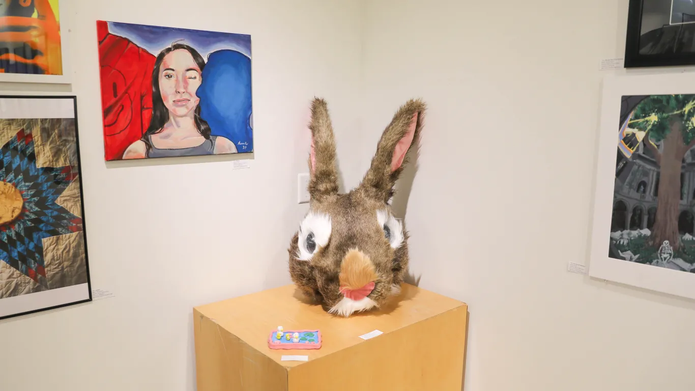 This is a group of art pieces on display in the Gallery consisting of acrylic paintings, photographs, colored pencil illustrations, a small duck pond made of clay and most notably, a large rabbit head made of paper mache and fake fur. 