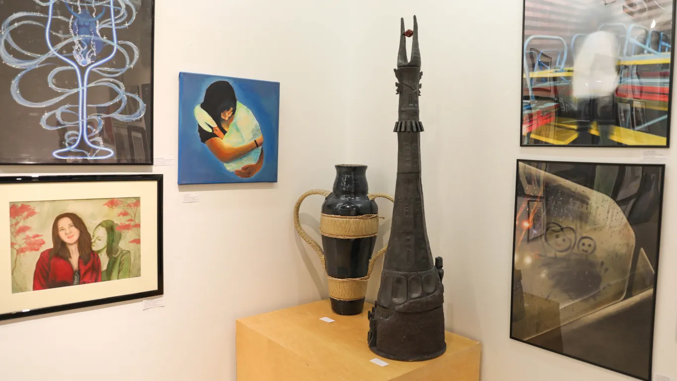 This is a group of art pieces on display in the Gallery consisting of mixed media illustrations, acrylic paintings, photographs and most notably, two large ceramic art pieces, a vase and a cinematic tower made of clay. 