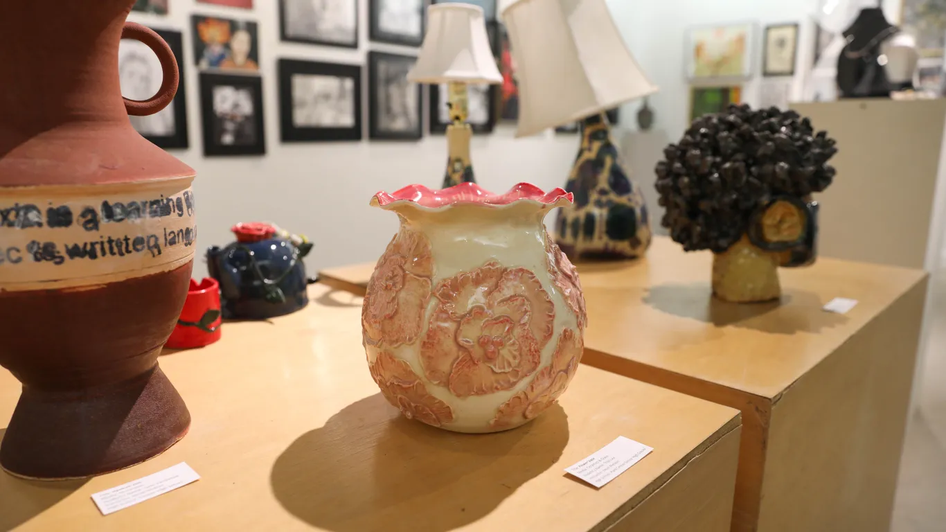 This is a group of ceramic art pieces on display in the Gallery. The flower vase has red, pink and white flowers on it and a wavy, shell-like, pink glazed edge. 