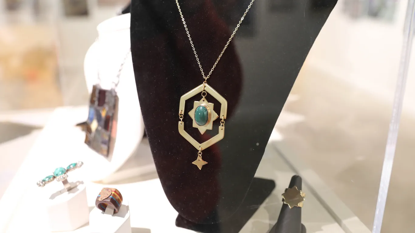 This is a group of jewelry designed by high school students on display in the Gallery. The pendant necklace consists of a gold chain with a green stone in the center. 