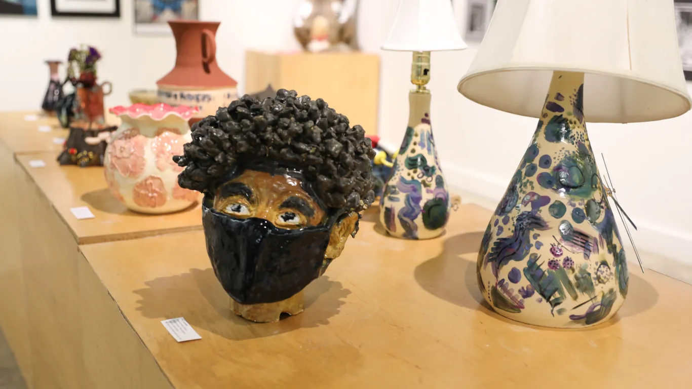 This is a group of ceramic pieces on display in the Gallery. There are two matching ceramic lamp designs and a ceramic head of a boy with an afro hairstyle, who is wearing a mask. 