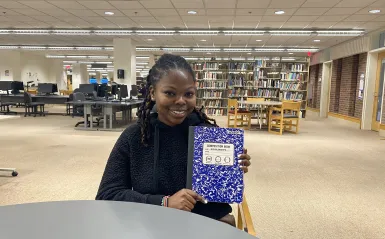 this is a photo of NHCC student, Lizzie, sitting in the library, posing with her blue composition notebook that she filled up with scholarships.