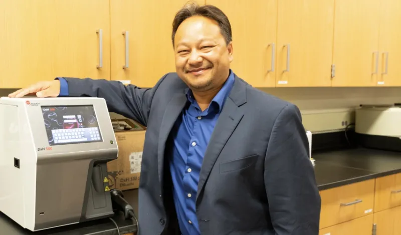 alumni Chue Yang standing over the wo hematology analyzers that were donated by Beckman Coulter Incorporated the company he now works for