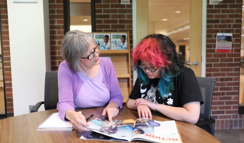 NHCC Workforce Innovation and Experiential Learning Center Director, Karen Philbin, sits with a female student at a table in the library looking at a career magazine together