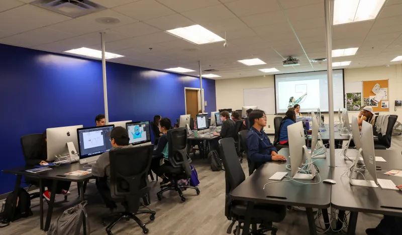 this is a photo of a graphic design classroom in NHCC's Fine Arts Center. Students are working at the iMac desktop computer stations, sitting in comfortable chairs, the teacher is pointing at a graphic on the screen at the front of the room, the wall is bright, dark blue and the vinyl floor is updated.