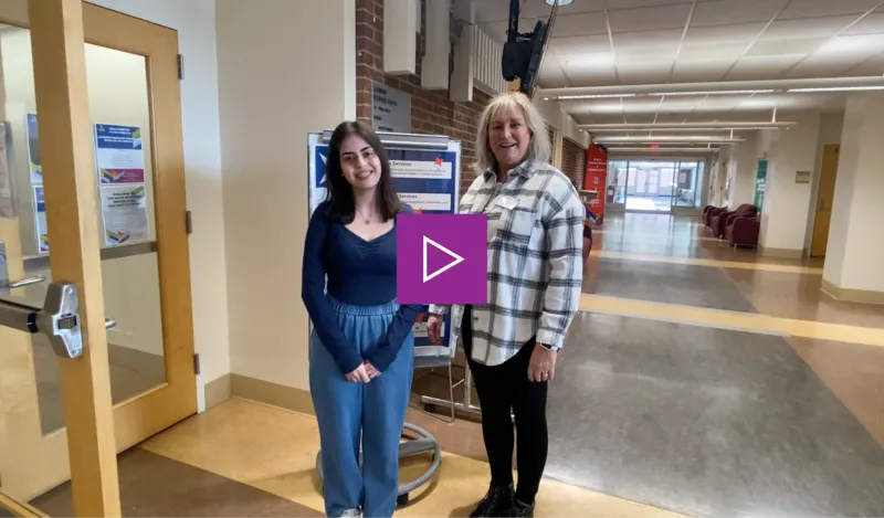 a photo of NHCC staff, Debi Long, with a student worker smiling standing in the hallway outside of the Library on-campus.