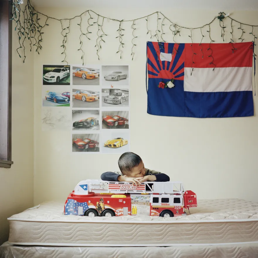 a photo of a little boy and his fire truck toy laying on his bed, underneath a flag of the Karen State