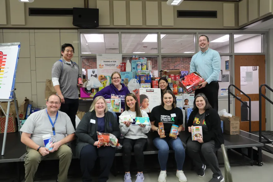 a group photo of NHCC staff smiling with food donations in the Campus Center during their Food Drive event. 