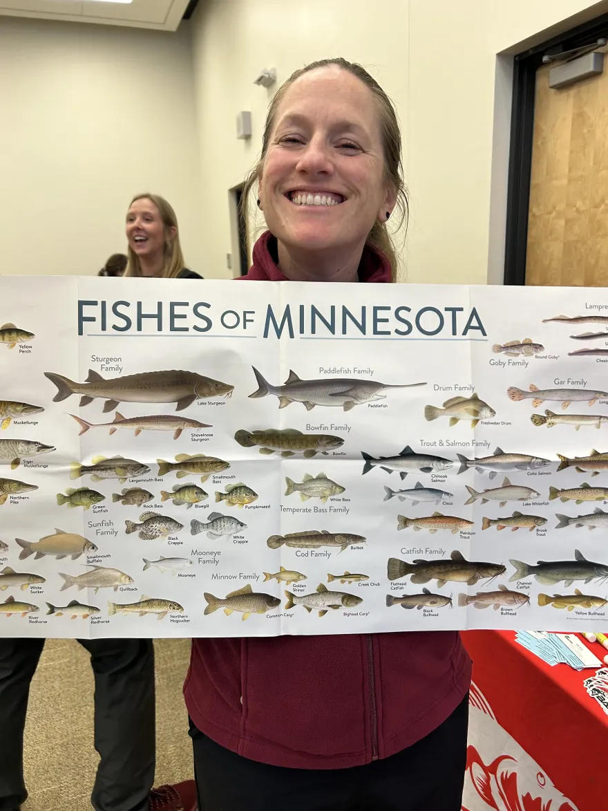 a photo of NHCC employee, Christie, smiling at the Green Jobs Fair event with a Fish of Minnesota poster on-campus in Helling Hall. 