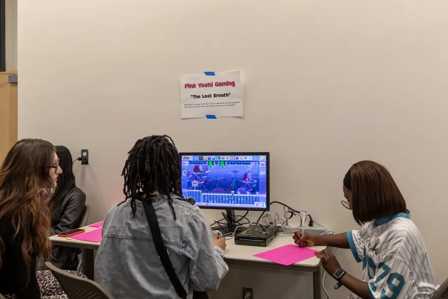 a photo of a female student playing a video game with two other female bystanders watching nearby. 