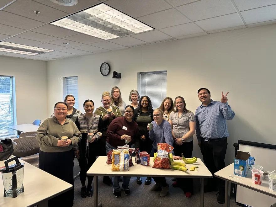 a group photo of NHCC employees gathering next to a table of frozen fruit and other ingredients for a Smoothie Party on-campus celebrating health and wellness goals. 