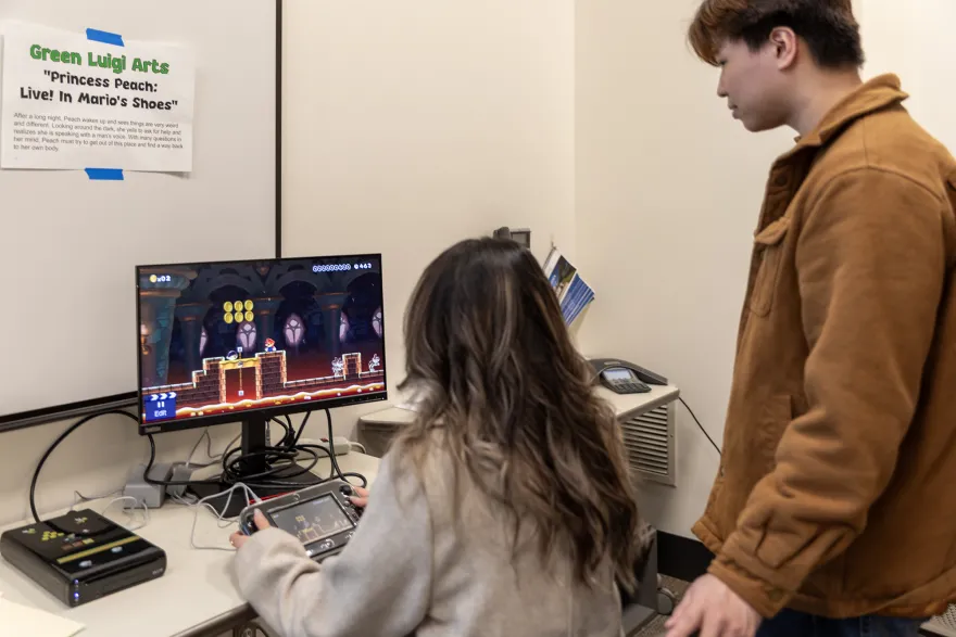 a photo of a female student playing and testing a student designed video game and a male student viewing the game behind her. 